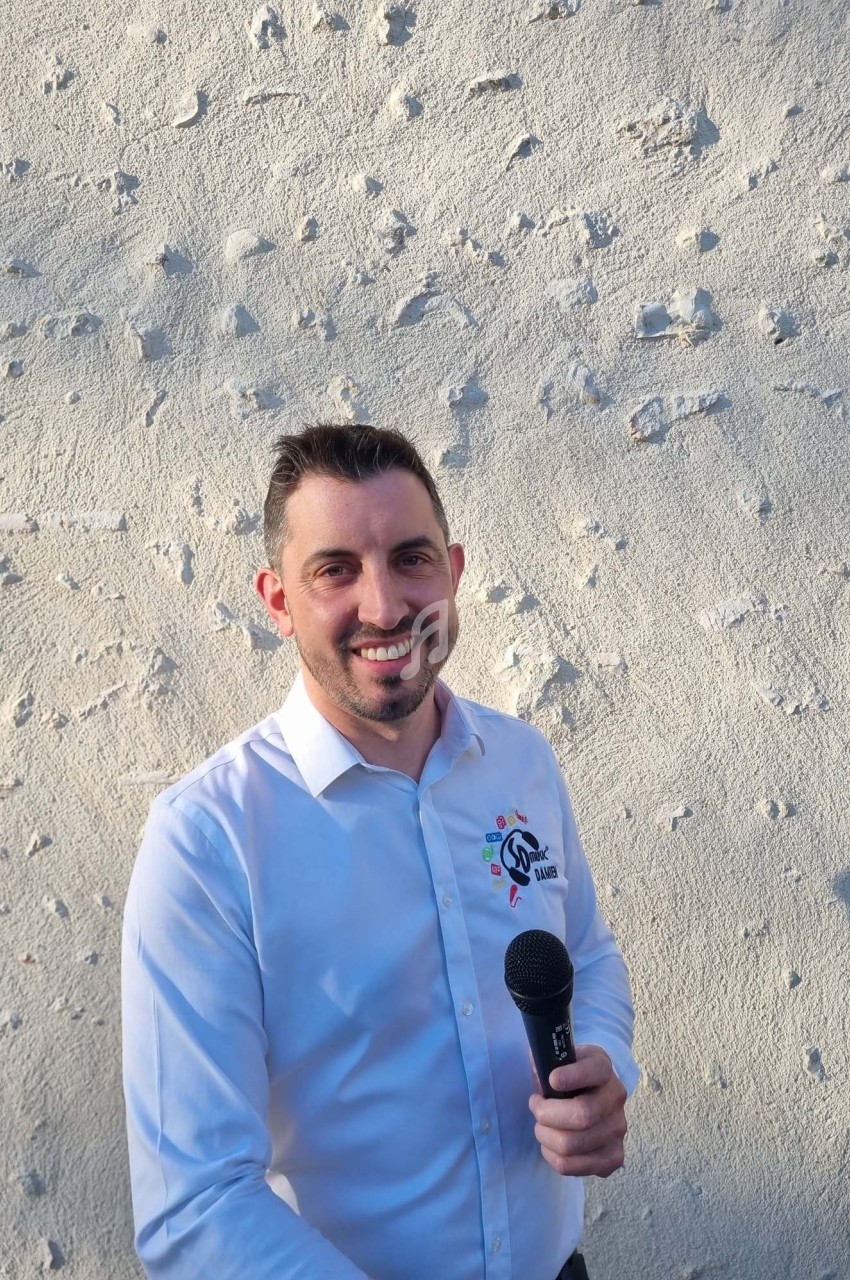 Homme souriant en chemise blanche tenant un micro devant un mur clair texturé.