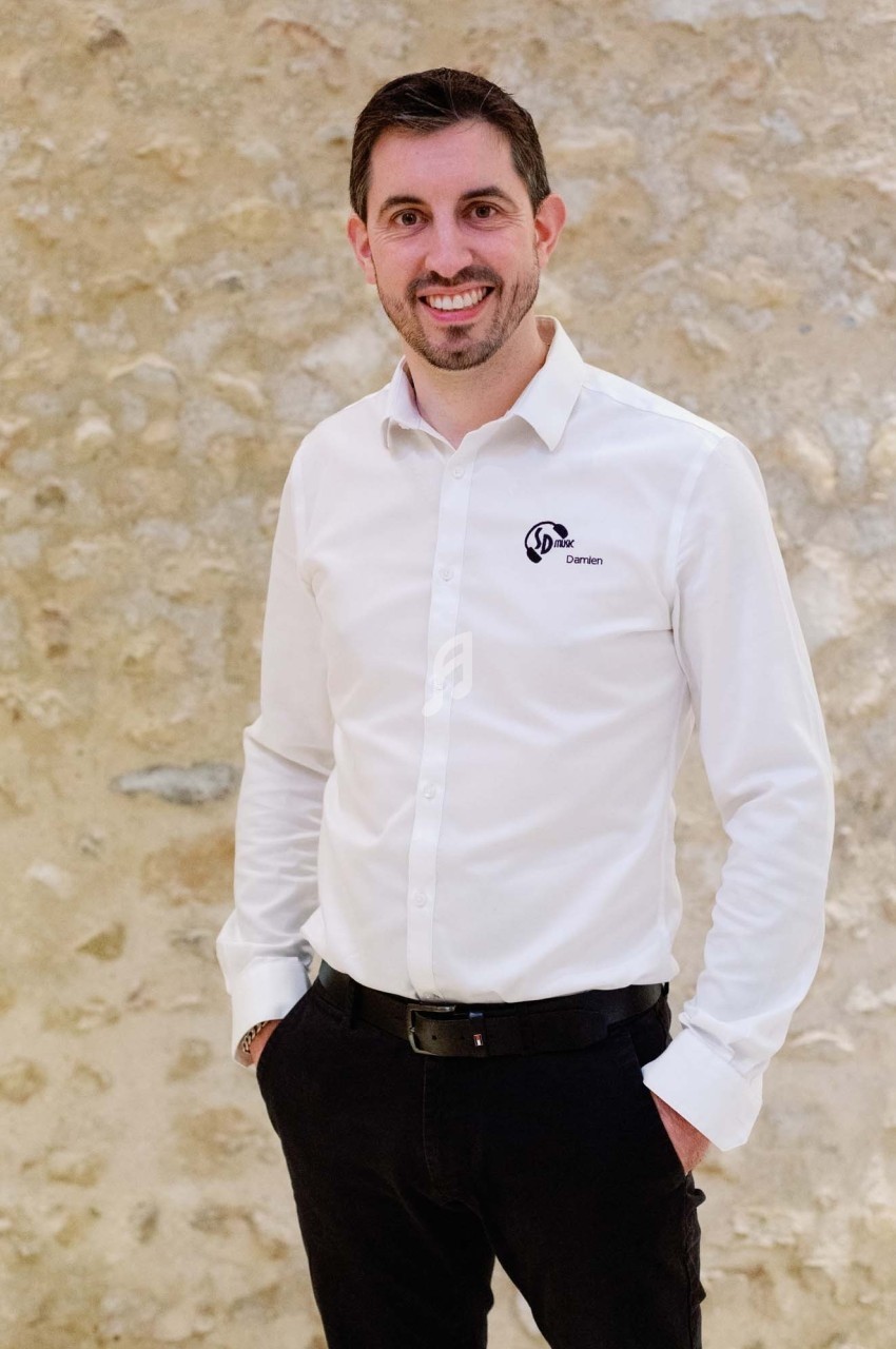 Homme souriant en chemise blanche et pantalon noir, debout devant un mur en pierre beige.