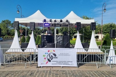 Scène extérieure avec un DJ sous des tentes blanches, entourée de barrières et équipée de matériel d'éclairage.