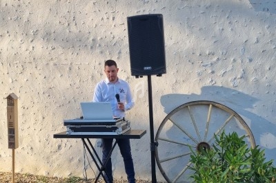 Un homme utilise un ordinateur portable et un micro à côté d'une enceinte, devant un mur extérieur avec une roue décorative.