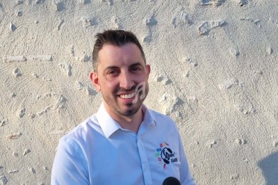 Homme souriant en chemise blanche tenant un micro devant un mur texturé clair.