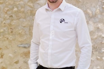 Homme souriant en chemise blanche et pantalon noir, debout devant un mur en pierre beige.