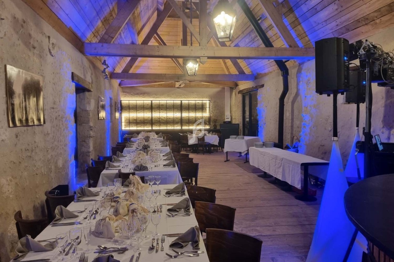 Salle de réception rustique avec poutres apparentes, tables dressées pour un repas et éclairage tamisé bleu.