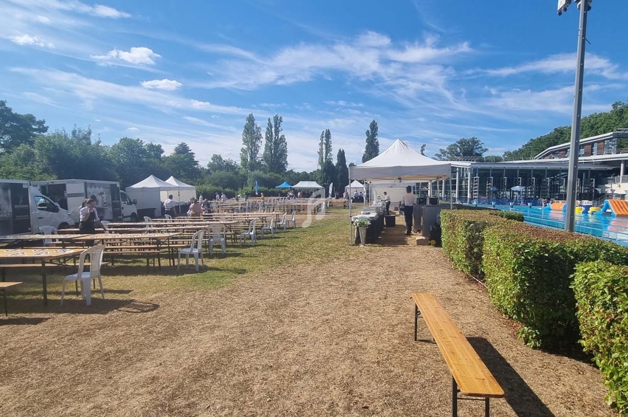 Des tables et bancs en bois sont disposés sur une pelouse près d'une piscine extérieure, sous un ciel dégagé.