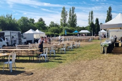Tables et chaises disposées en extérieur sur une pelouse, avec des stands, des tentes et des camions en arrière-plan.