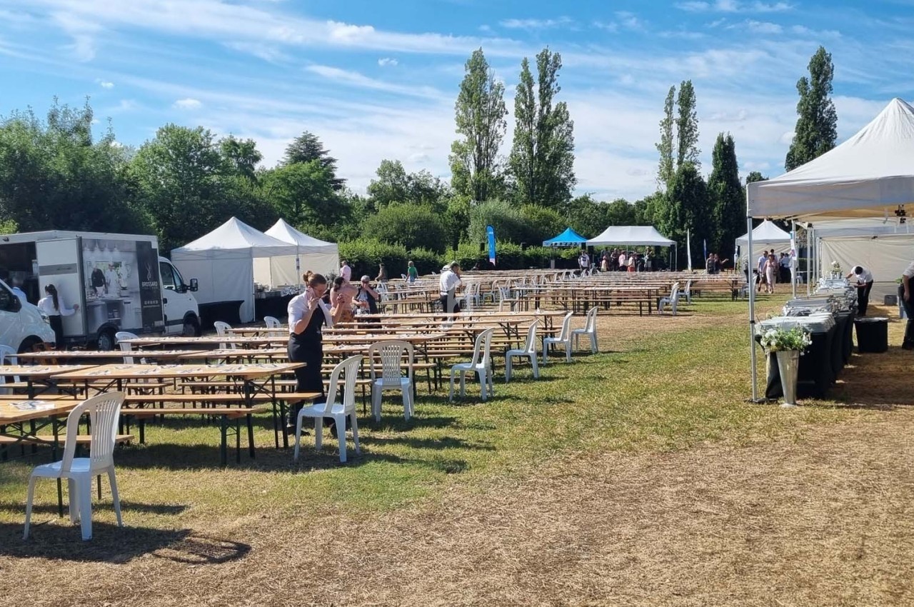 Tables et chaises disposées en extérieur sur une pelouse, avec des stands, des tentes et des camions en arrière-plan.