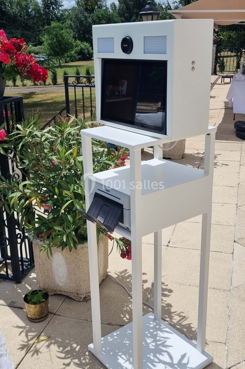 Borne photo blanche sur pied avec écran et imprimante intégrée, installée en extérieur sur une terrasse ensoleillée.