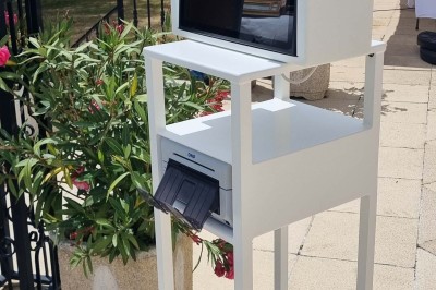 Borne photo blanche sur pied avec écran et imprimante intégrée, installée en extérieur sur une terrasse ensoleillée.