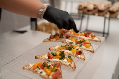 Une main gantée dispose des toasts garnis de fromage frais, tomates et herbes sur un plateau transparent.
