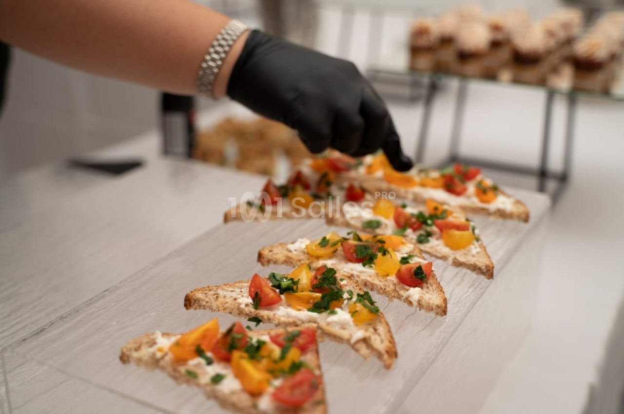 Une main gantée dispose des toasts garnis de fromage frais, tomates et herbes sur un plateau transparent.