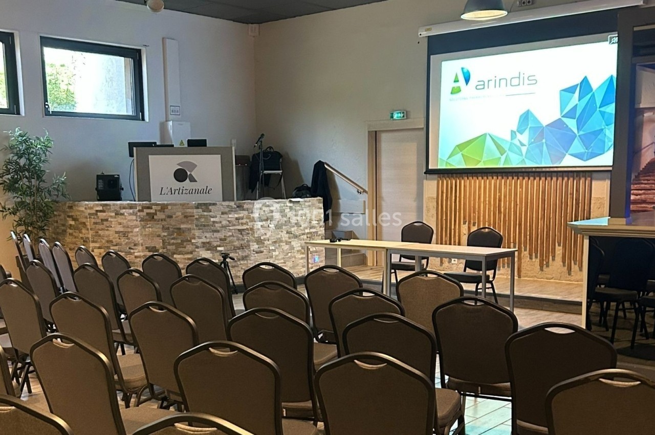 Salle de conférence avec chaises alignées, écran de projection allumé et pupitre sur une estrade en pierre.