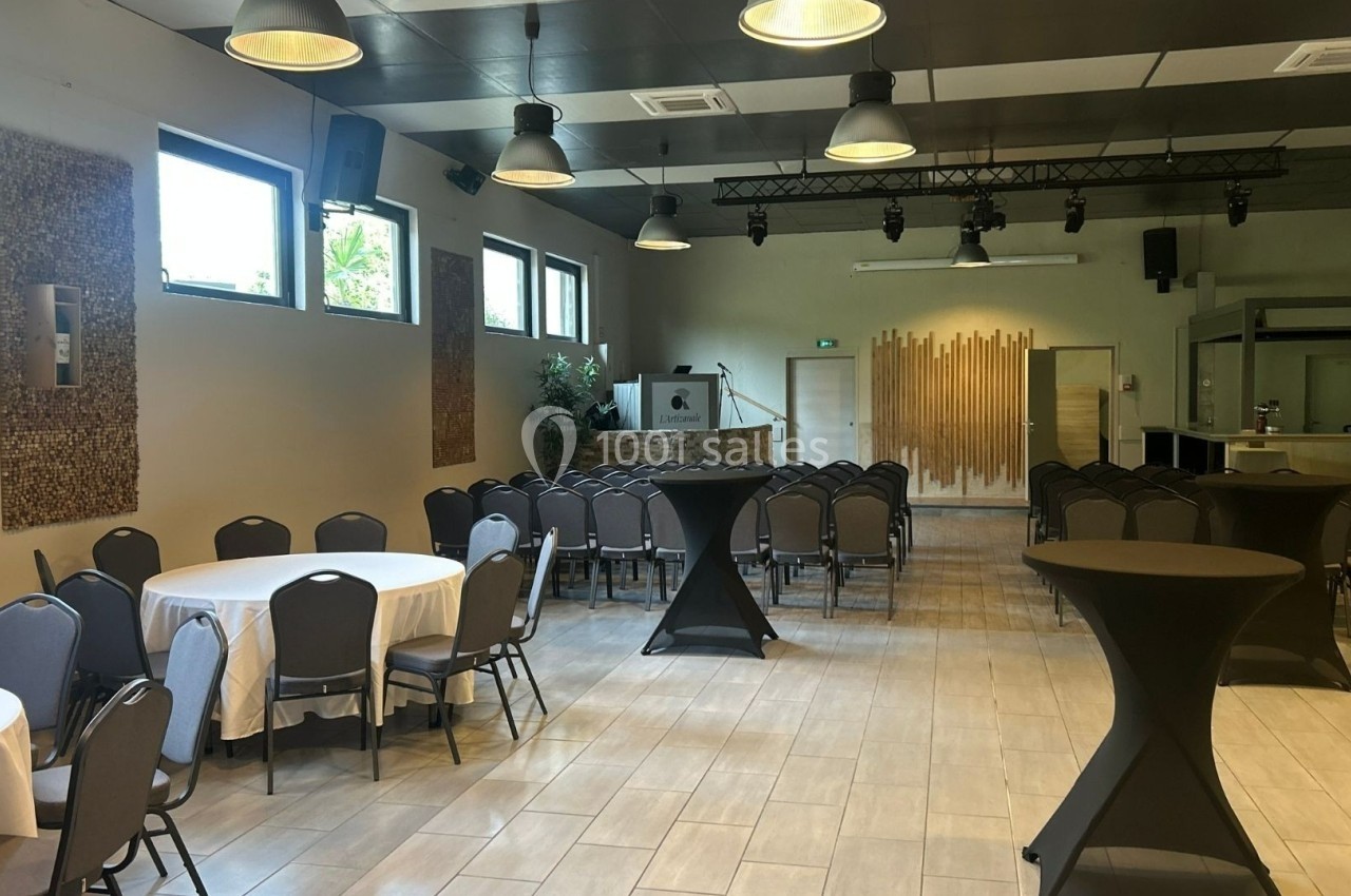 Salle de réception lumineuse avec tables rondes dressées, chaises alignées et un espace scénique en arrière-plan.