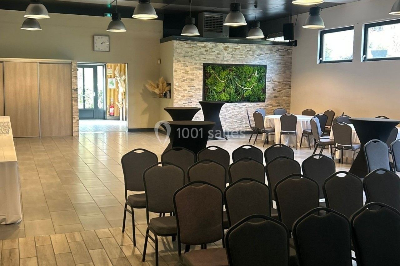 Salle lumineuse aménagée pour un événement, avec chaises alignées, tables rondes dressées et décoration végétale murale.
