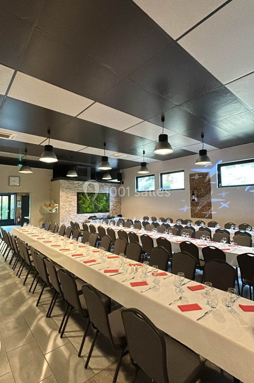 Salle de réception avec de longues tables dressées, nappes blanches, serviettes rouges et chaises alignées.