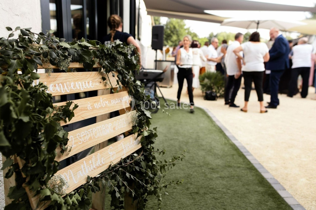 Panneau en bois décoré de feuillage annonçant un mariage, avec des invités rassemblés en arrière-plan.