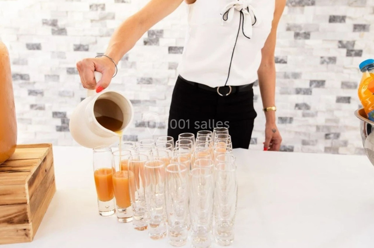 Une personne verse un jus orange dans des verres alignés sur une table blanche, avec un fond de mur en pierre claire.