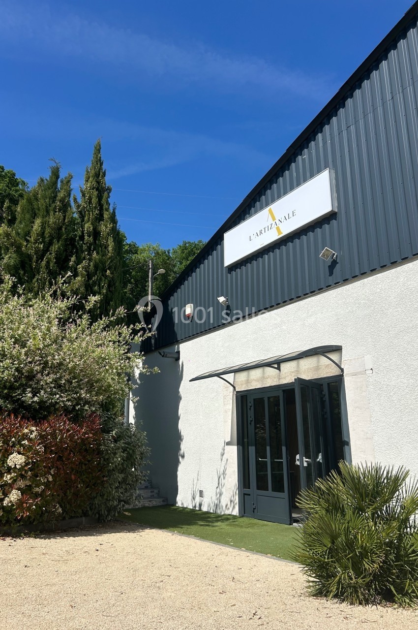 Façade d'un bâtiment moderne avec un panneau ’L'Artisanale’, entouré de végétation sous un ciel bleu.
