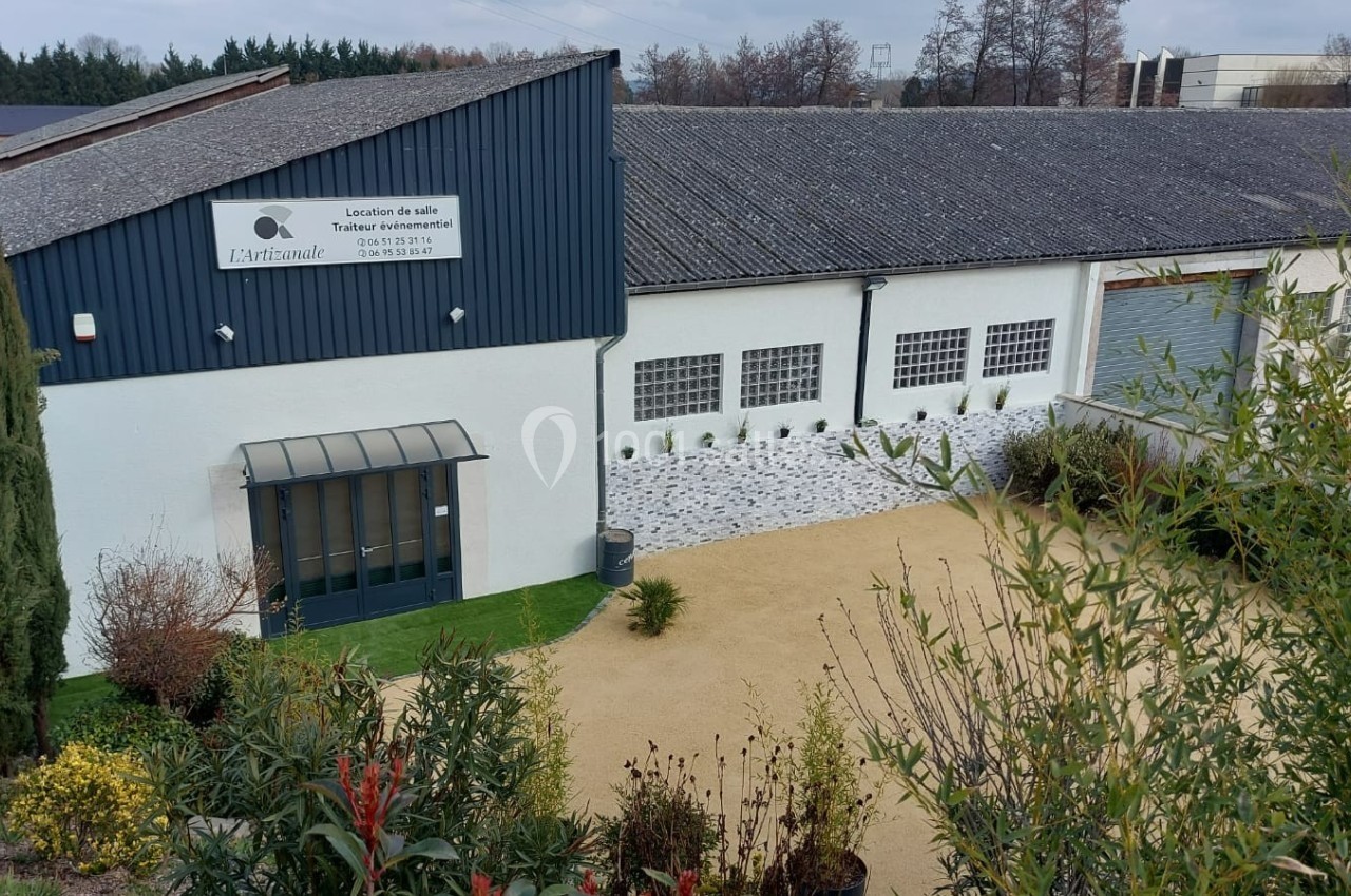 Bâtiment avec façade blanche, entrée vitrée, panneau signalant une location de salle, entouré de végétation.