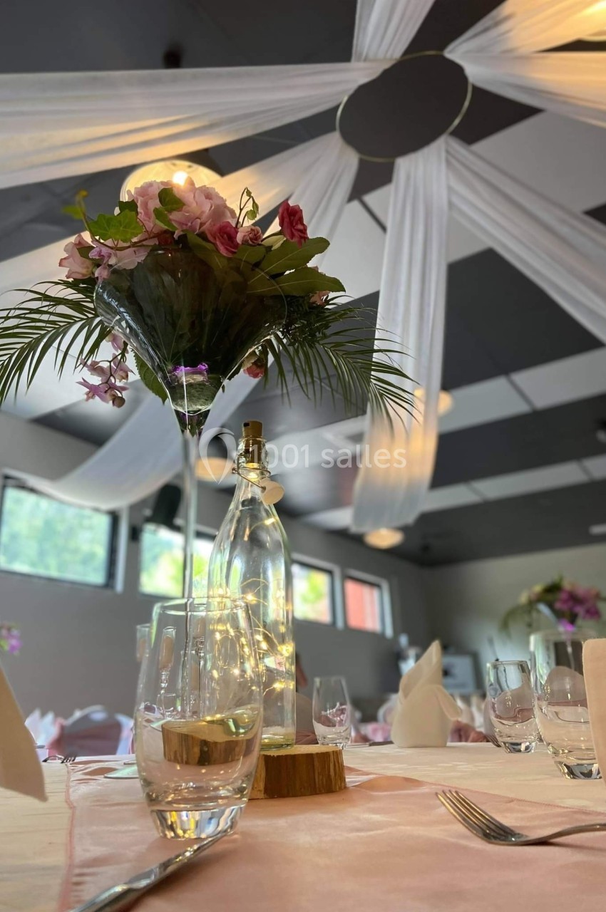 Centre de table avec fleurs, feuillage, bouteille lumineuse et nappes blanches dans une salle décorée pour un événement.
