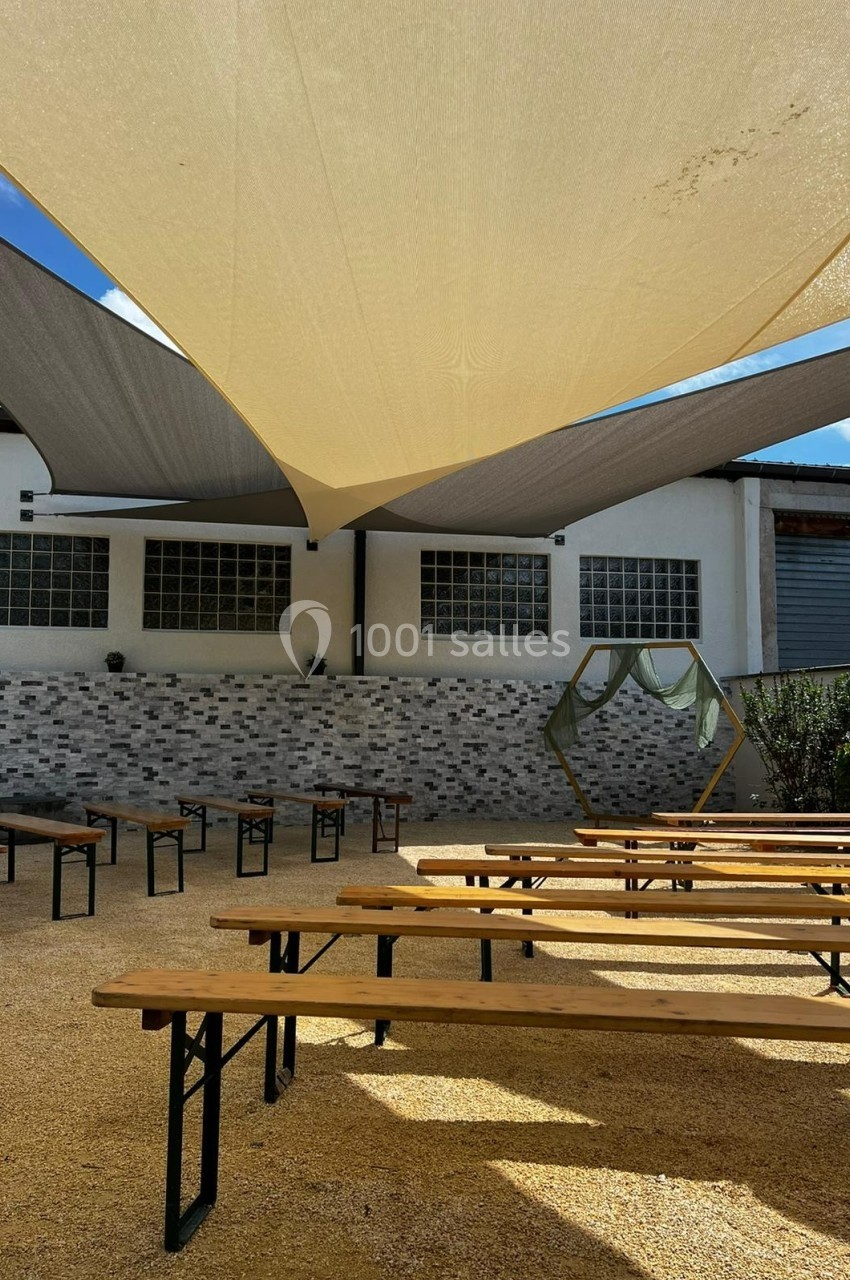 Bancs en bois disposés sur une terrasse en gravier, ombragée par des voiles tendues devant un bâtiment industriel.