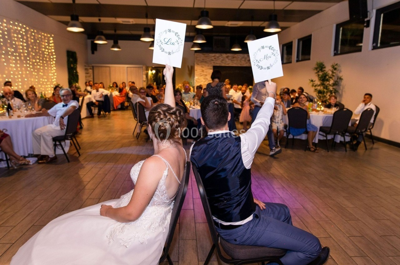 Un couple assis dos à dos lors d'un jeu de mariage, chacun levant une pancarte dans une salle décorée.