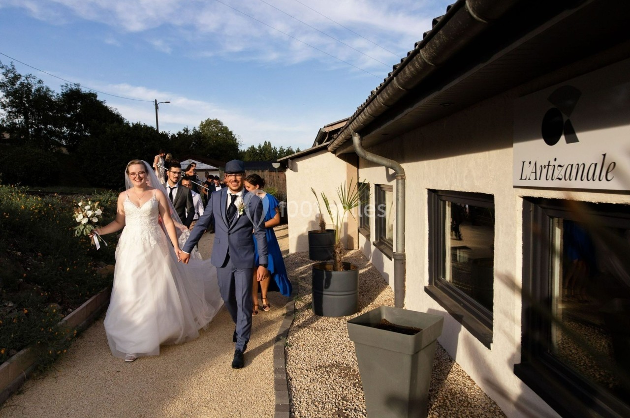 Un couple de mariés marche main dans la main, suivi de leurs invités, près d'un bâtiment nommé ’L'Artizanale’.