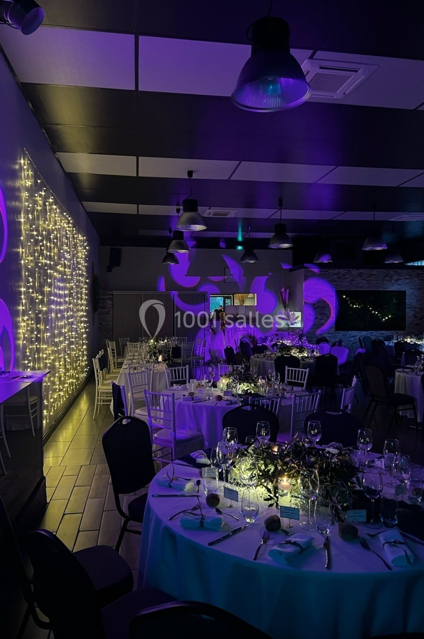 Salle de réception décorée avec des guirlandes lumineuses, des tables dressées et un éclairage tamisé violet.