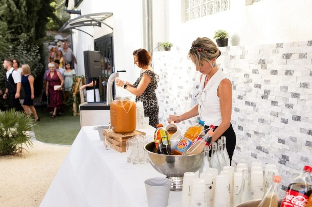Deux femmes préparent un buffet de boissons en extérieur, avec des invités en arrière-plan près d'un jardin.
