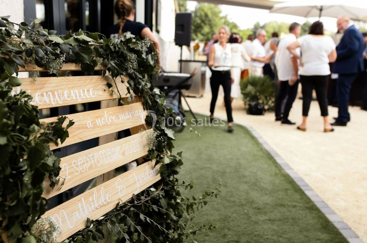 Panneau en bois décoré de feuillage annonçant un mariage, avec des invités rassemblés en arrière-plan extérieur.
