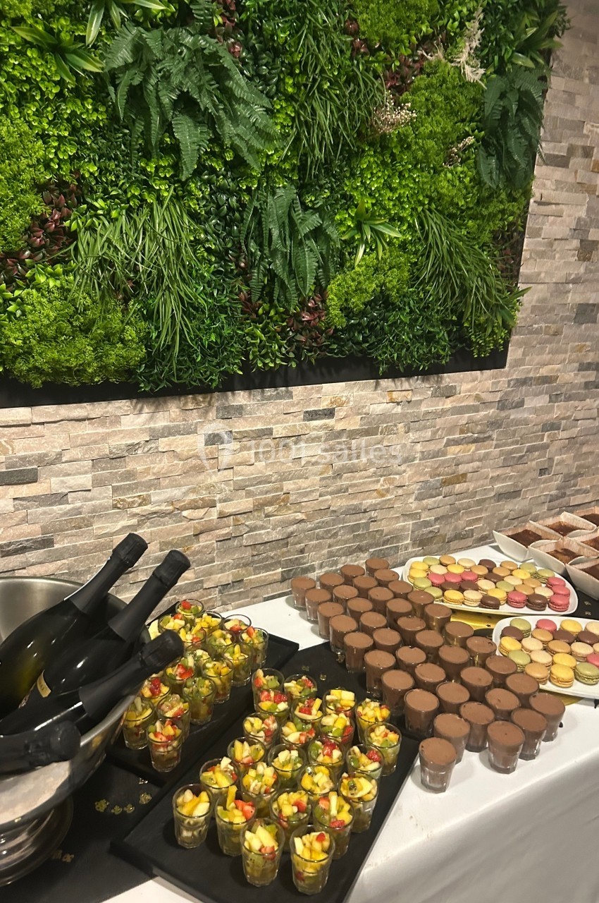 Buffet avec desserts variés, verrines de fruits, macarons et mousse au chocolat, devant un mur végétal décoratif.