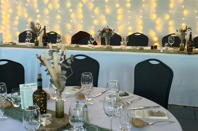 Table de réception décorée avec nappes blanches, verres, couverts, centres de table végétaux et guirlandes lumineuses en…
