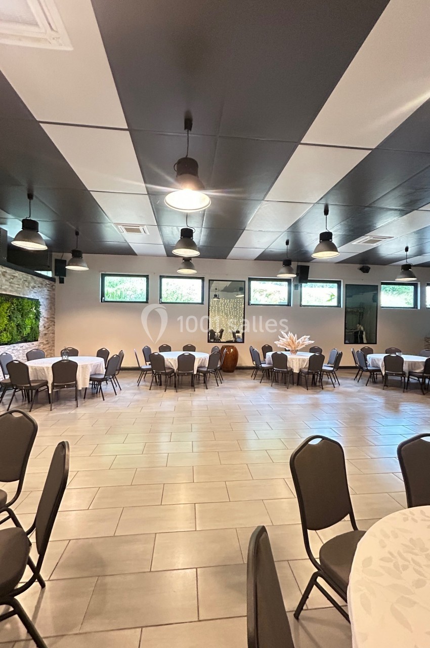 Salle spacieuse avec tables rondes dressées, chaises noires, murs en pierre et grandes fenêtres laissant entrer la lumière.