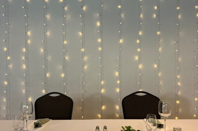 Table de réception décorée avec nappes blanches, verres, couverts, centres de table végétaux et guirlandes lumineuses en…