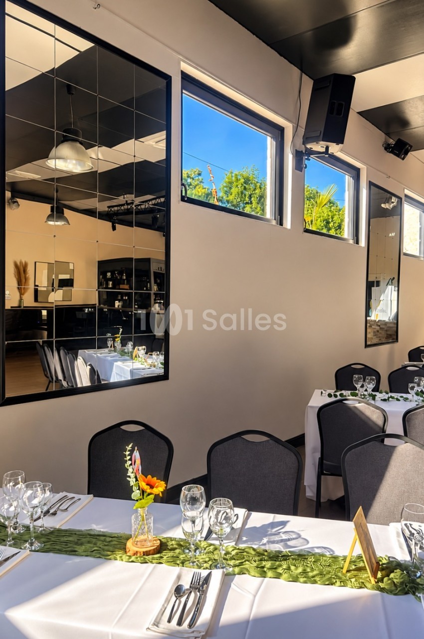 Salle de réception aux tables dressées de nappes blanches, chemins de table verts et centres floraux, chaises noires et grands miroirs muraux.