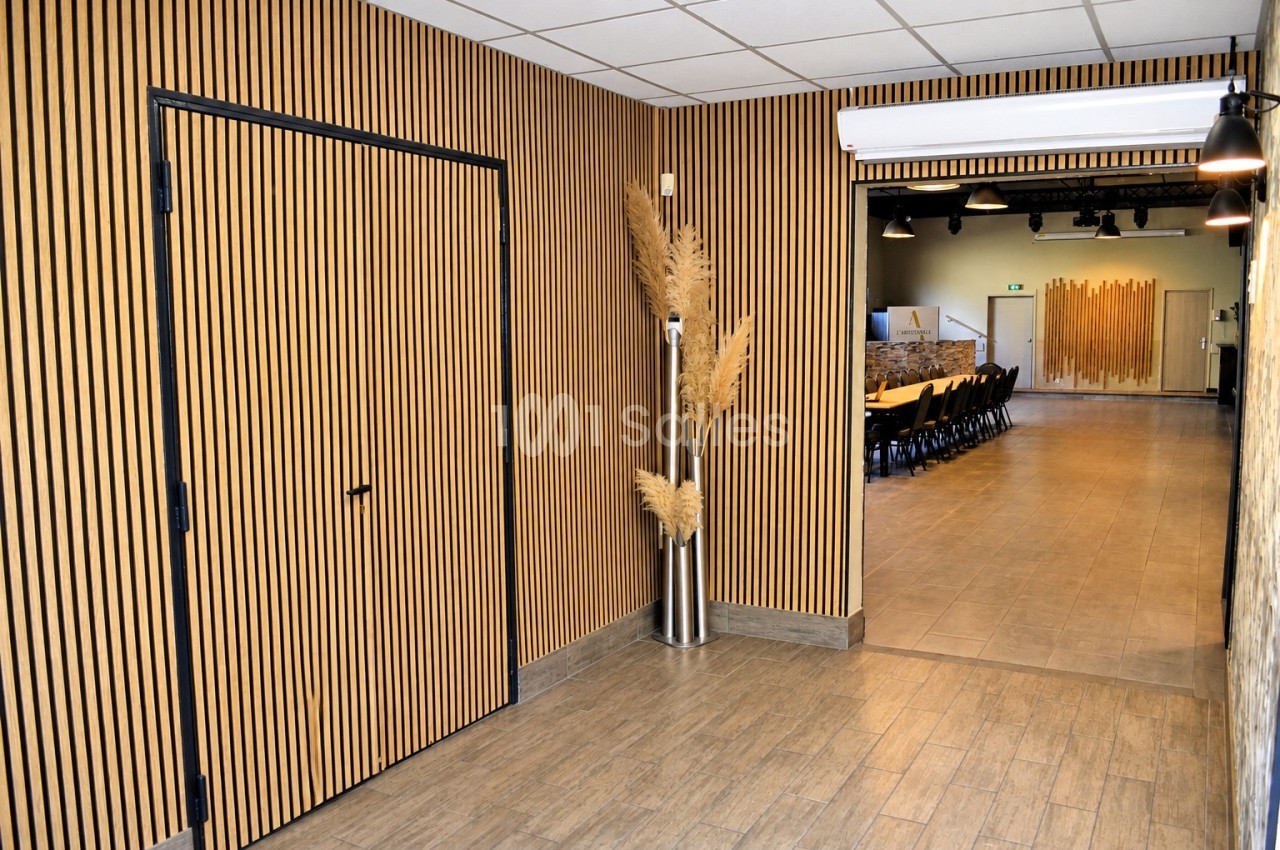 Couloir aux murs en lattes de bois clair menant à une salle de réception dressée de tables et chaises, avec sol carrelé imitation bois.