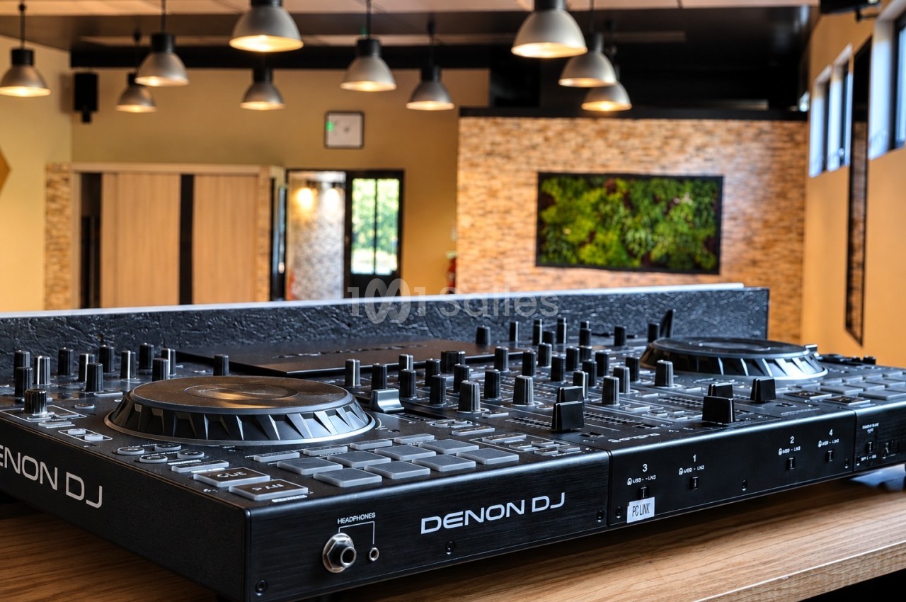Console de mixage DJ noire posée sur une table en bois, dans une salle éclairée de suspensions et mur végétalisé.