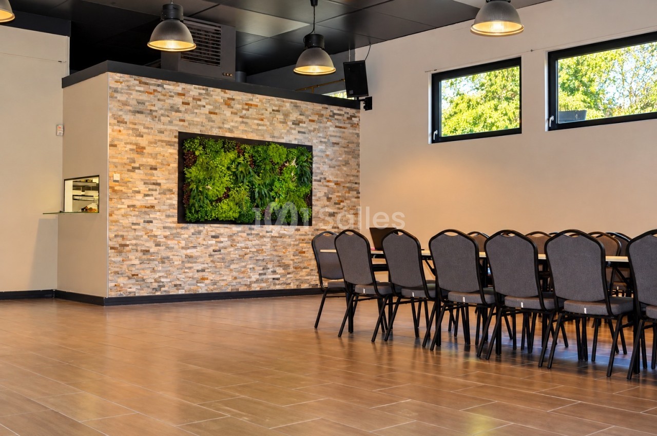 Salle aux murs de pierre et d'un mur végétalisé, avec rangées de chaises grises alignées le long d'une table, sol en carrelage imitation bois.