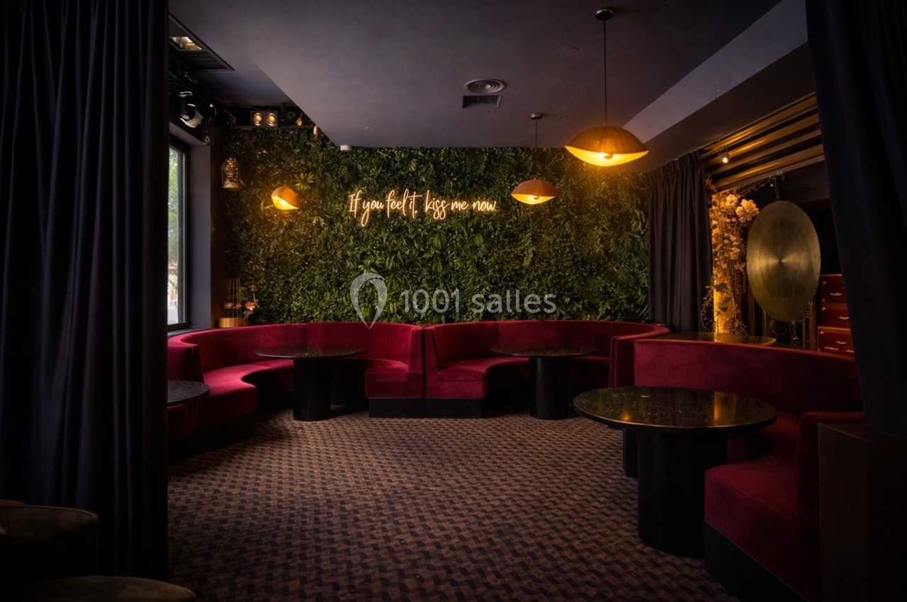 Salon avec banquettes rouges, tables basses noires, mur végétal éclairé par une enseigne lumineuse et lumière tamisée.