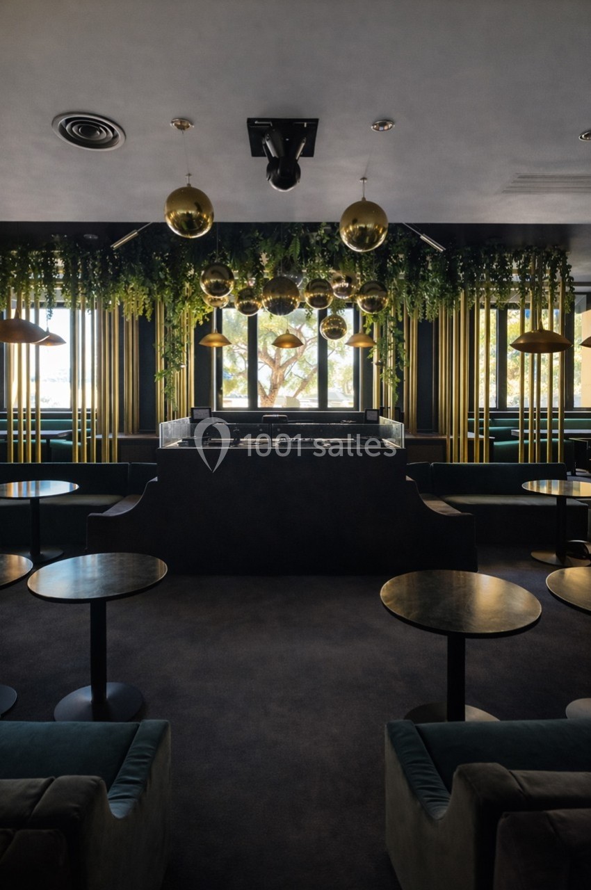 Salle moderne avec tables rondes, banquettes vertes, luminaires dorés suspendus et plantes décoratives au plafond.