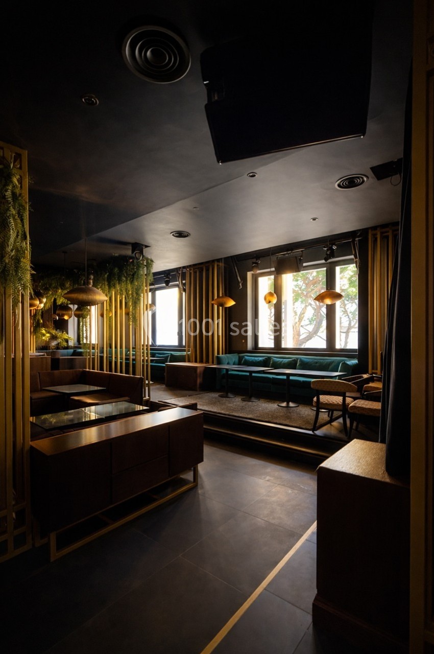 Salle intérieure sombre avec mobilier moderne, luminaires suspendus, plantes décoratives et grandes fenêtres.