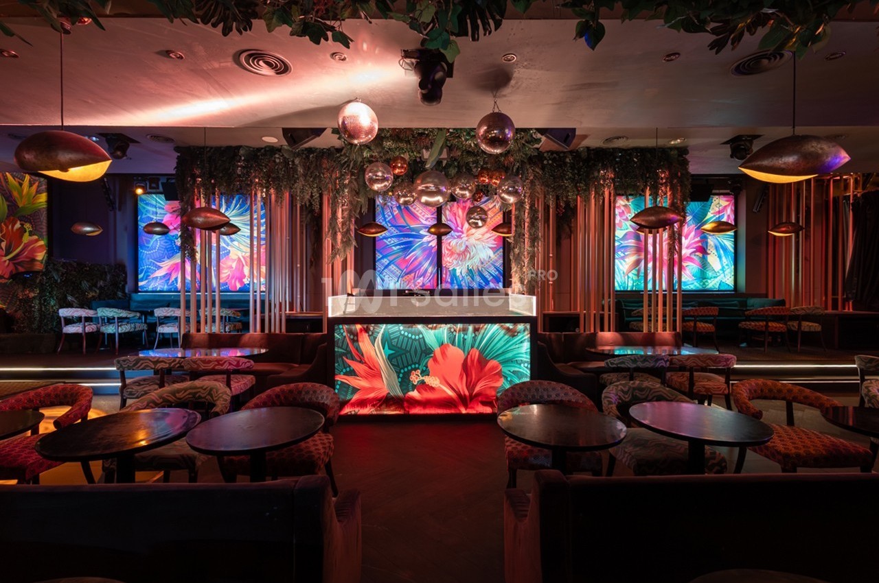 Salle de bar colorée avec décorations florales, éclairage tamisé et écrans affichant des motifs lumineux.
