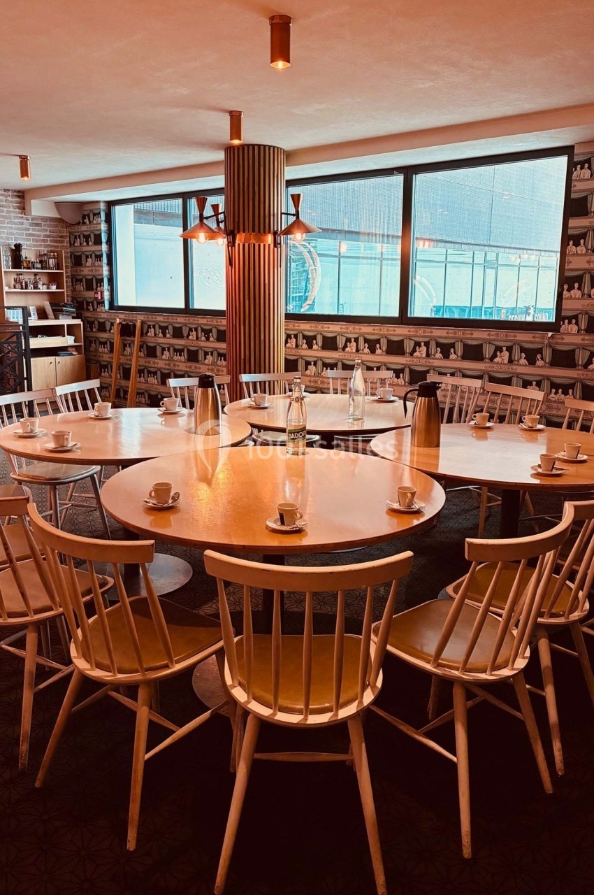Salle avec une grande table ronde en bois entourée de chaises, éclairée par des suspensions et une lumière naturelle.