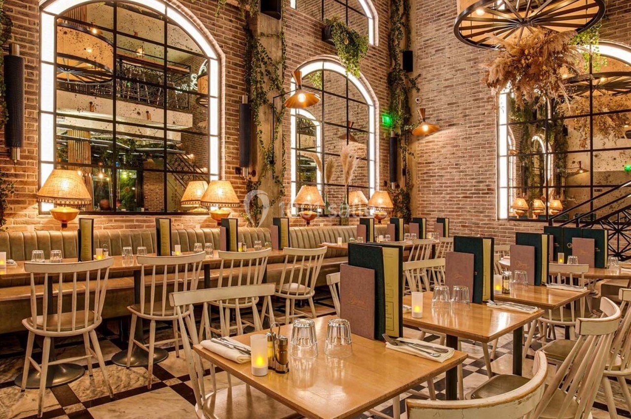Salle de restaurant chaleureuse avec murs en briques, grandes fenêtres, tables en bois et éclairage tamisé.