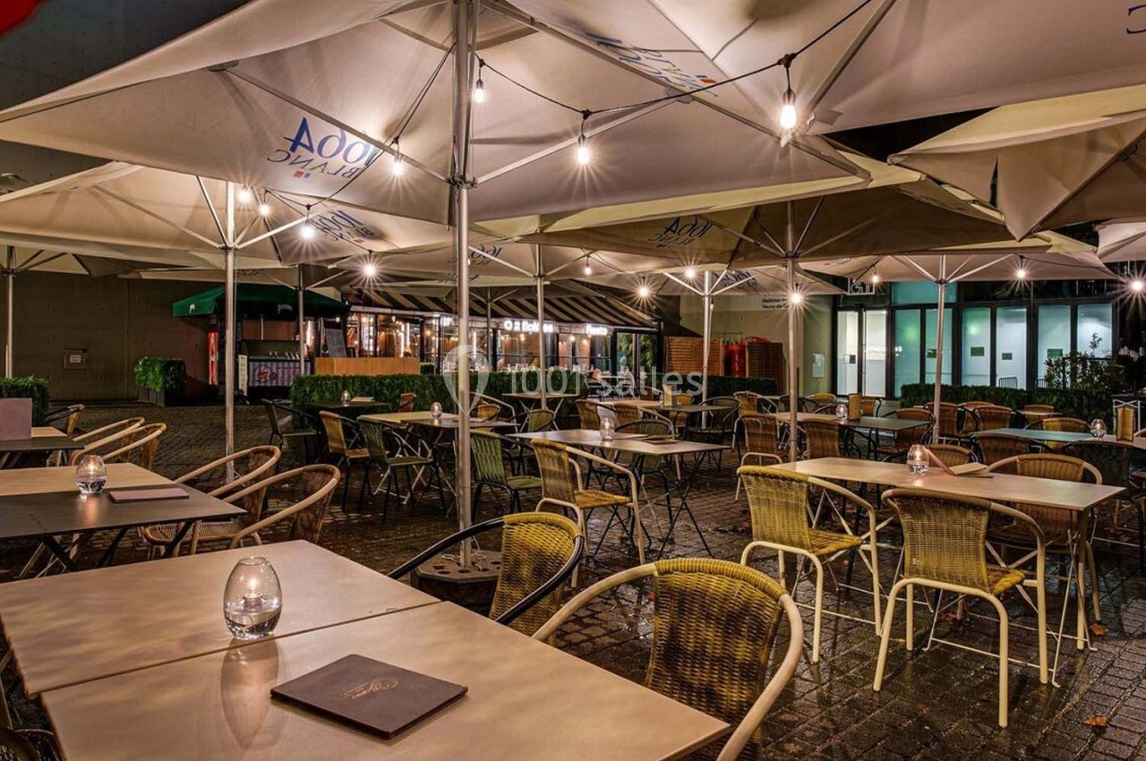Terrasse extérieure d'un restaurant éclairée en soirée, avec des tables et des chaises sous de grands parasols.