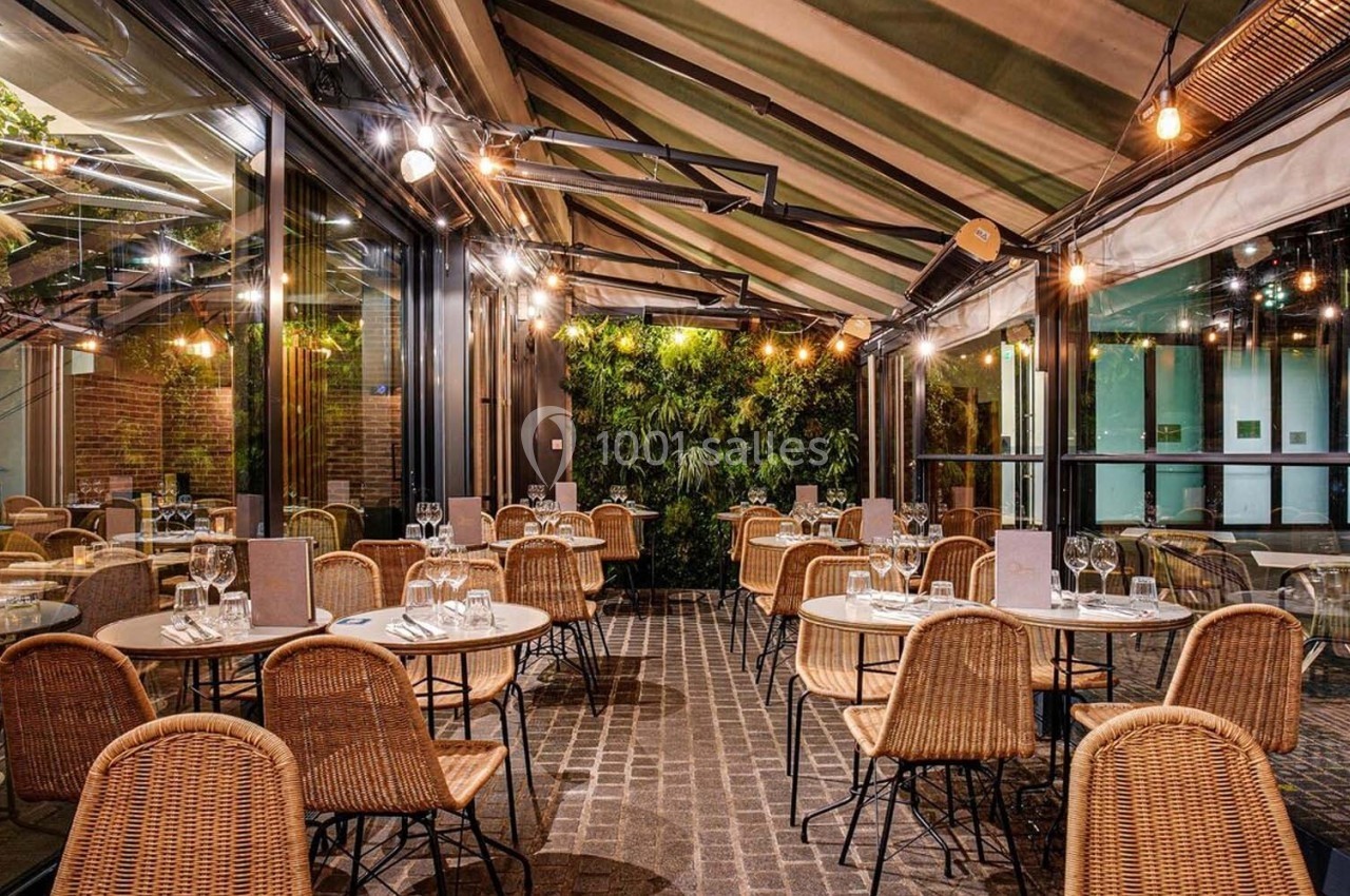 Terrasse couverte avec tables rondes, chaises en rotin, éclairage chaleureux et mur végétal en arrière-plan.