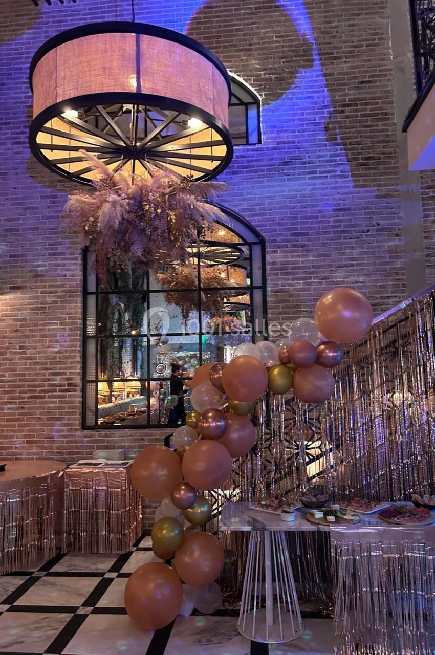 Décoration festive avec guirlande de ballons roses et dorés, pampas suspendues et mur en briques dans un espace éclairé.