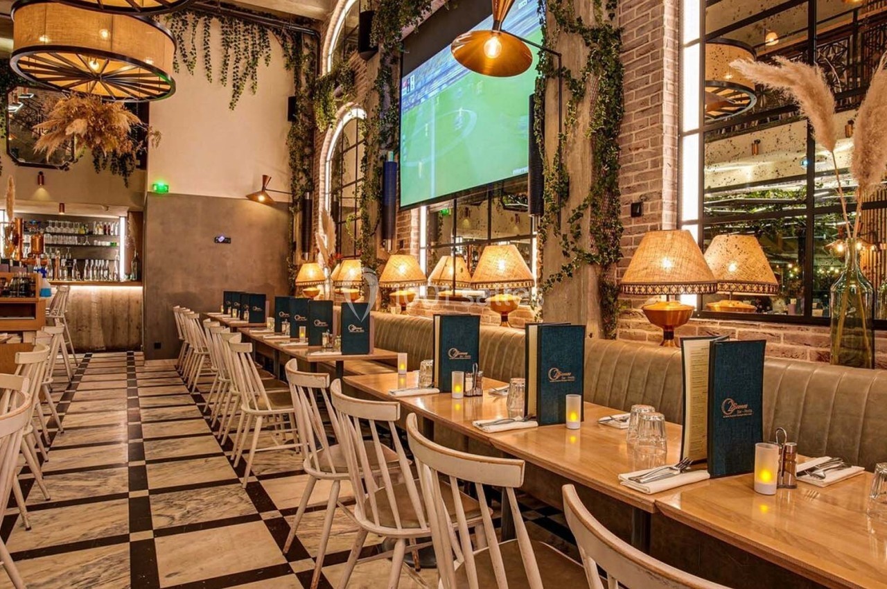 Salle de restaurant chaleureuse avec tables alignées, chaises en bois, éclairage tamisé et décoration végétale.