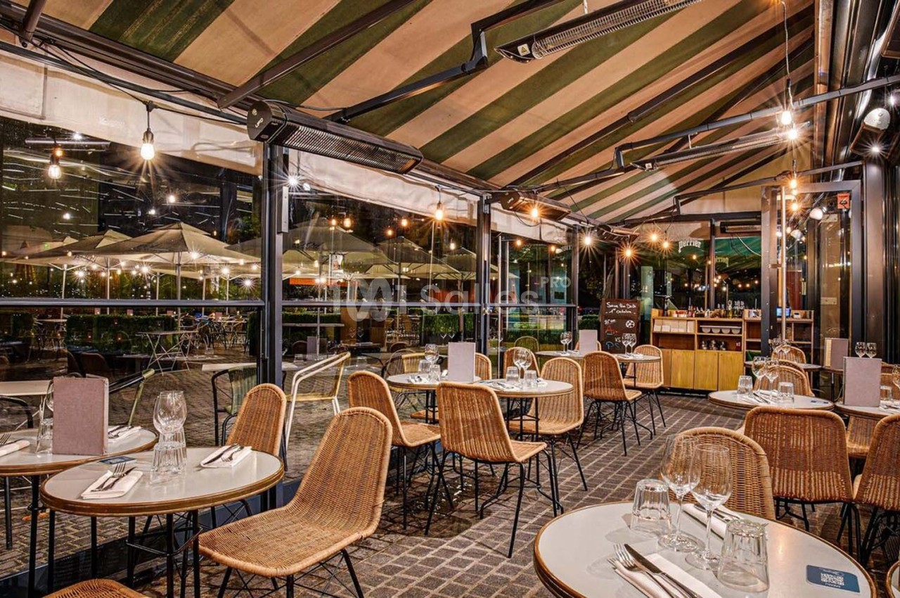 Terrasse couverte d'un restaurant avec tables dressées, chaises en rotin et vue sur un extérieur éclairé en soirée.