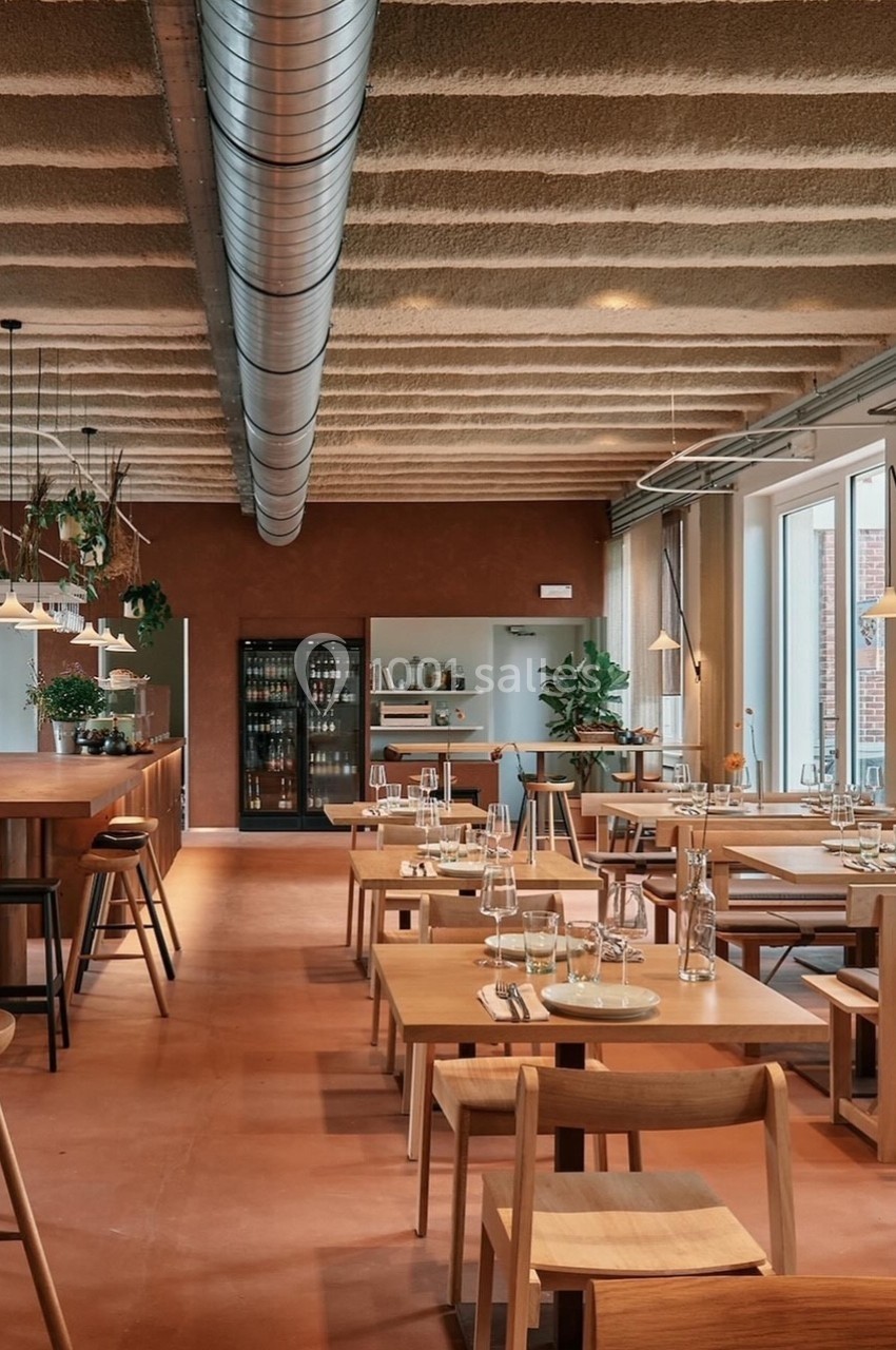 Salle de restaurant moderne avec tables en bois, grandes fenêtres, éclairage suspendu et décoration végétale.