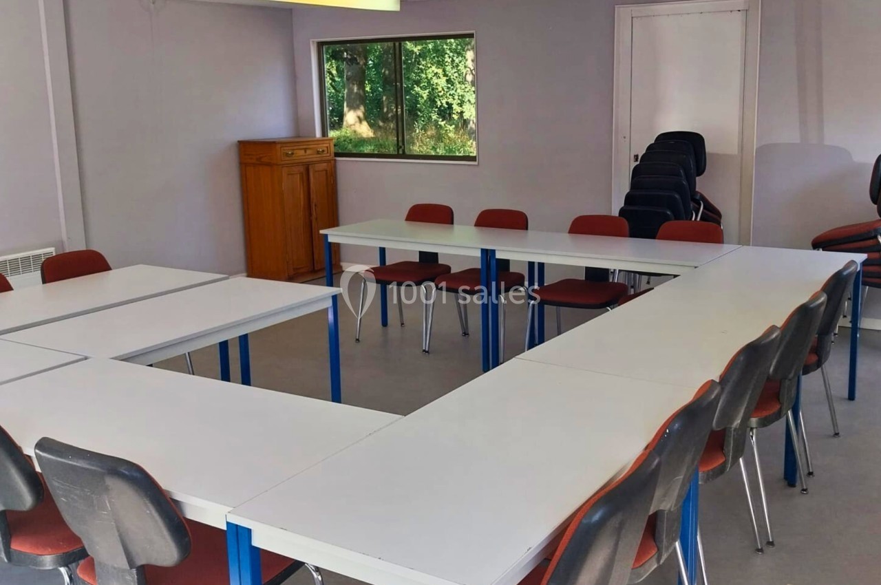 Salle de réunion lumineuse avec des tables disposées en U, entourées de chaises rouges et vue sur un espace extérieur.